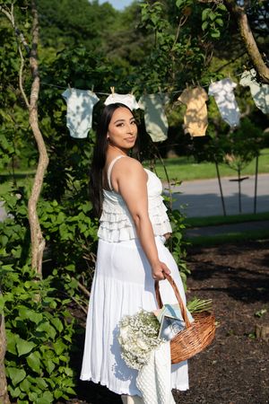 A pregnant woman in a white dress holds a wicker basket with flowers in a sunny garden setting.