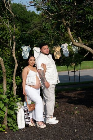 A pregnant woman and her partner pose together in white outfits under baby clothes hanging on a line.
