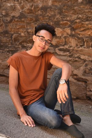 Young man in rust orange t-shirt and jeans sitting against a stone wall outdoors.
