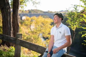 Young man in white polo and green cap sits on wooden fence surrounded by lush trees and scenic bridge view.