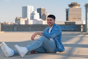 Laughing young man in denim jacket sits on rooftop ground with urban city skyline behind him.