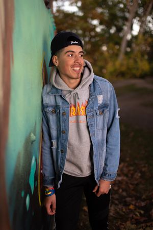 Smiling teen in denim jacket and black cap leans against colorful graffiti wall in autumn setting.