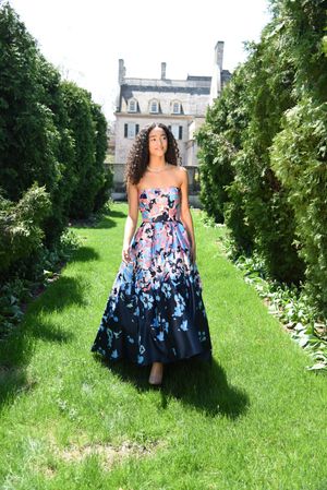 Woman in strapless floral ball gown walking through a garden path with a historic mansion backdrop.
