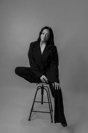Black and white studio portrait of woman in black suit seated on stool in elegant pose.