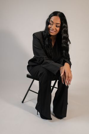 Woman with long black hair in black pantsuit smiling while seated on folding chair in studio.