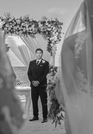Groom standing at beach wedding altar under floral arch, awaiting bride in black and white photo.