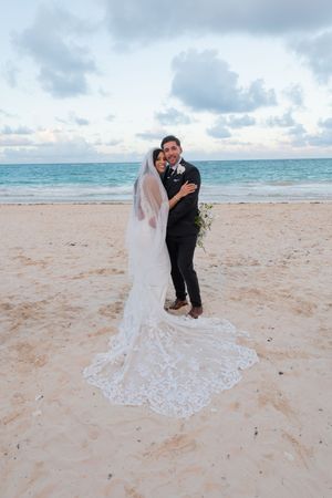 Bride and groom embrace on sandy beach, bride wearing lace wedding gown with long train and veil, ocean backdrop.
