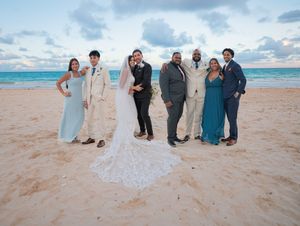 Wedding party of six poses on sandy beach at sunset, bride in lace gown with long veil at center.