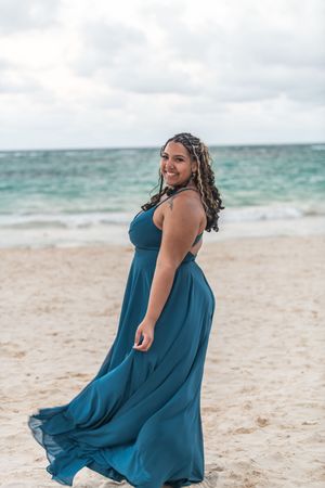 Woman in flowing teal gown poses on sandy beach with ocean waves behind her, smiling at camera.