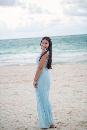 Young woman in light blue maxi dress stands barefoot on sandy beach, smiling over her shoulder.