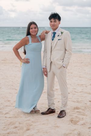 Couple poses on beach, woman in light blue bridesmaid dress beside man in beige suit with boutonniere.
