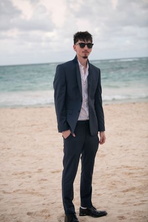 Stylish man in dark navy suit and sunglasses stands confidently on sandy beach with ocean behind him.
