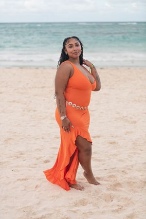 Woman in orange one-shoulder dress with chain belt poses barefoot on sandy beach by the ocean.