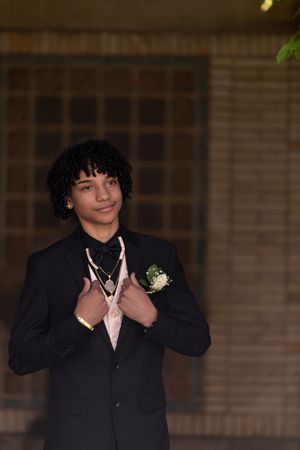 Stylish teen boy in black suit with boutonniere and pink tie poses confidently for prom photos.