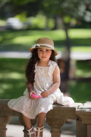 Cute young girl in floral dress and straw hat sits on stone railing in sunny park setting.