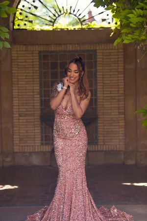 Young woman in rose gold sequin mermaid prom gown smiles in ornate brick archway setting.