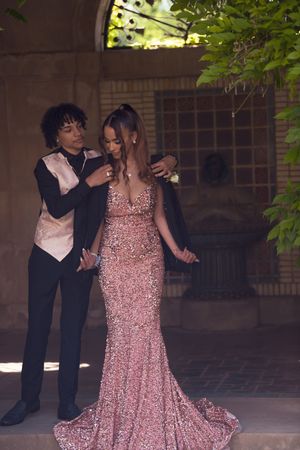 Teen prom couple in rose gold sequin gown and black tuxedo pose together in elegant brick archway.