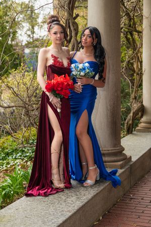 Two young women in formal red and blue gowns holding bouquets posing at an outdoor venue.