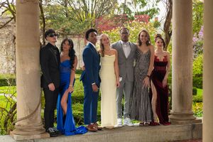 Group of seven prom-dressed teens posing together under stone columns in a garden setting.