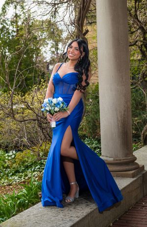 Young woman in royal blue mermaid prom gown holding blue floral bouquet beside a stone column.
