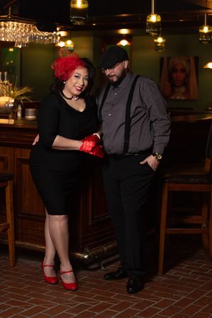 Stylish couple in retro outfits posing in an elegant bar, woman in black dress with red accessories.