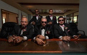 Group of five groomsmen in black tuxedos with pink bow ties posing at a bar counter.
