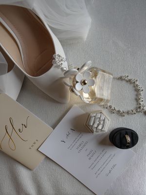 Flat lay of bridal accessories including white heels, vows card, wedding invitation, and jewelry.