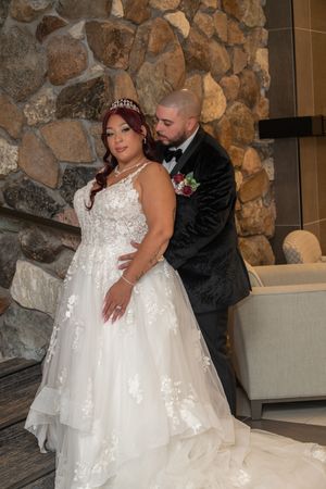 Bride in layered white lace gown with tiara and groom in black velvet suit by stone wall.