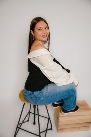 Young woman smiling in off-shoulder black and white top with blue jeans, seated on a stool against white background.