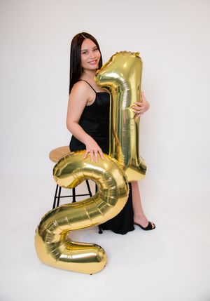 A smiling woman in a black dress poses with large gold number 21 balloons on a white background.