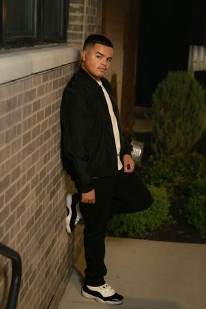 Young man in black bomber jacket leaning against brick wall at night wearing Jordan sneakers.