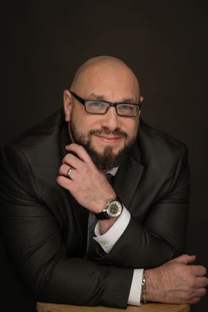 Bald bearded man in dark suit with glasses and watch posing confidently for professional headshot.
