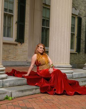 Young woman in stunning red velvet mermaid gown with gold bodice poses on steps by white columns.