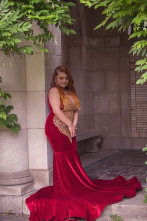 Young woman in elegant red velvet mermaid gown with gold lace bodice poses in stone archway.