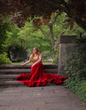 Young woman in flowing red velvet prom dress twirls on stone steps in lush green garden setting.