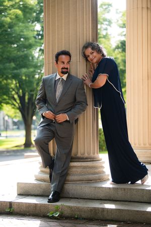 Elegant couple posing by stone columns outdoors, man in gray suit and woman in navy gown.