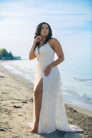 Elegant bride in white lace slit gown standing barefoot on a serene beach shoreline under blue sky.