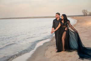 Couple in matching black outfits posing romantically on a sandy beach at golden hour sunset.