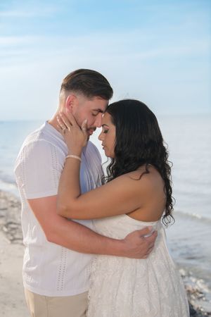 Romantic couple embracing on a sandy beach, touching foreheads tenderly with ocean and blue sky behind them.