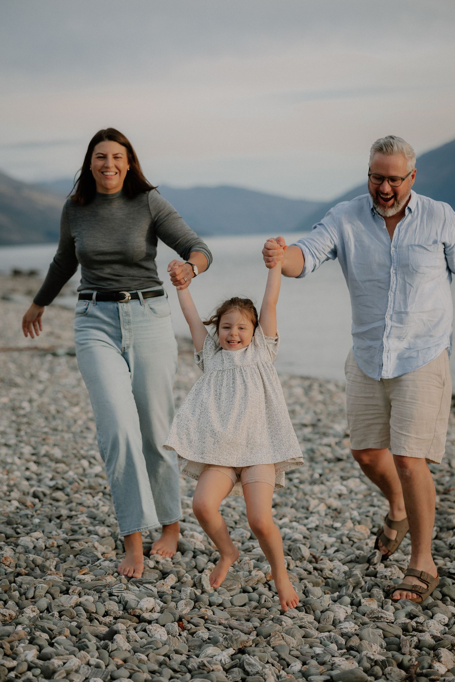 Family of three joyfully swinging young girl between parents on rocky beach with mountain lake backdrop.