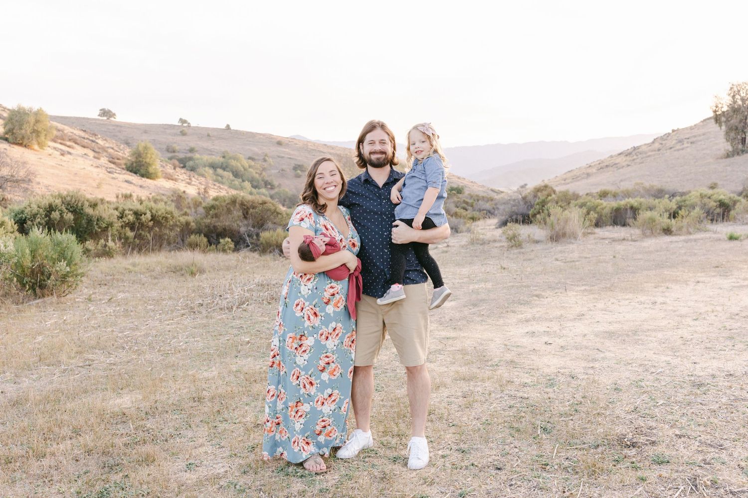 Happy family of three posing outdoors in a sunny field with rolling hills, parents holding toddler in casual summer attire.