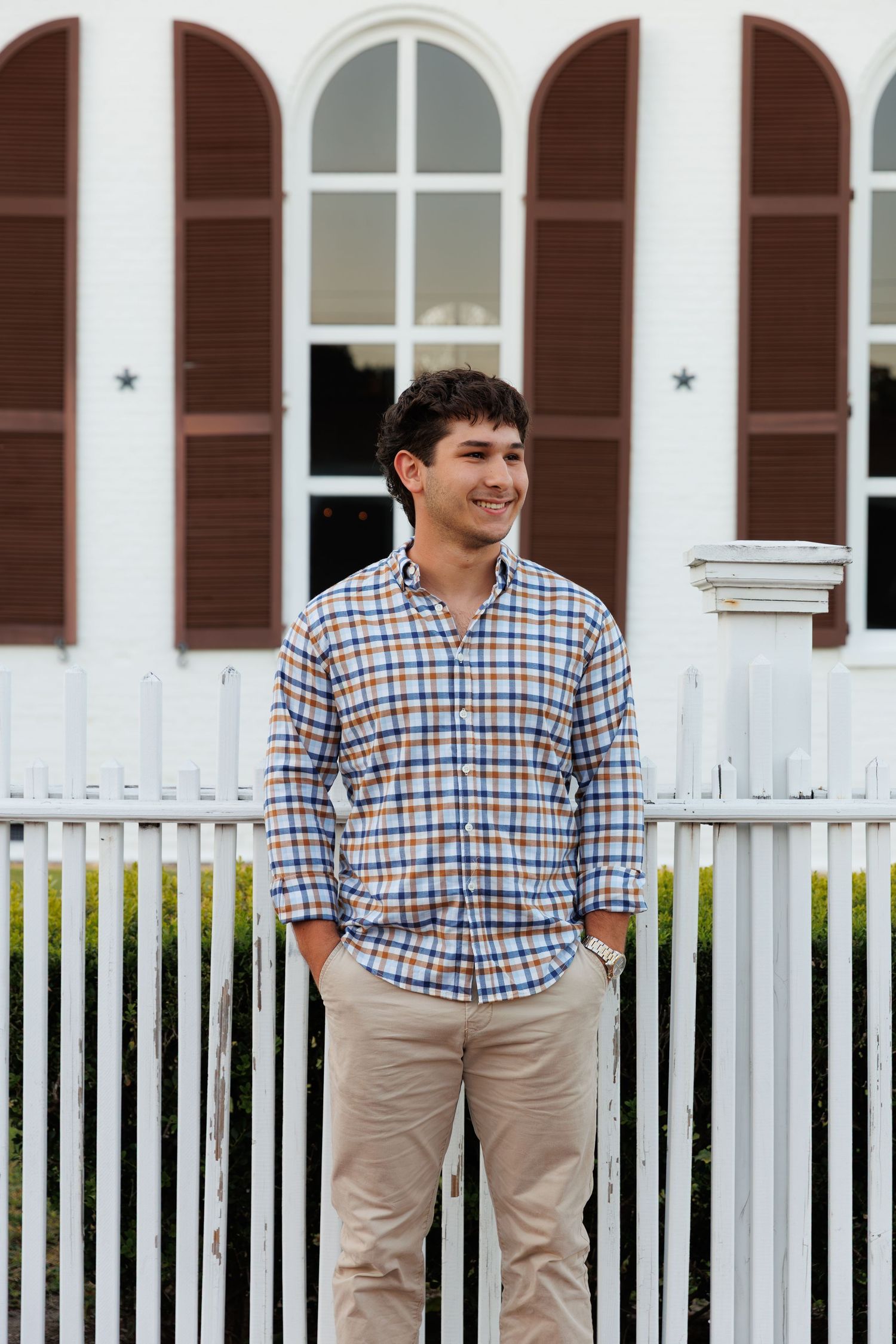 Young man smiling in plaid shirt and khakis standing in front of white picket fence and historic building.