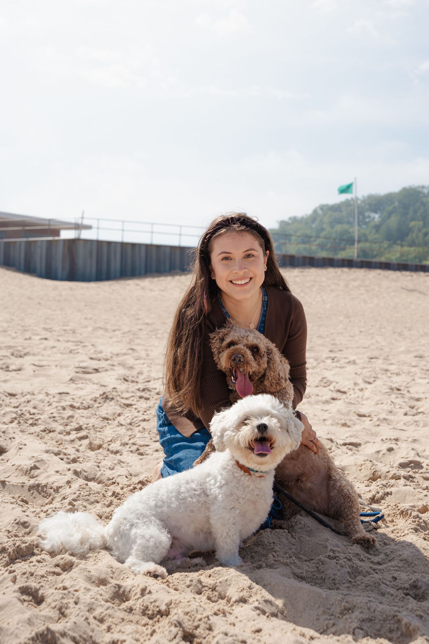 Smiling woman with long brown hair sitting on sandy beach with two fluffy dogs, white and brown, on a sunny day.