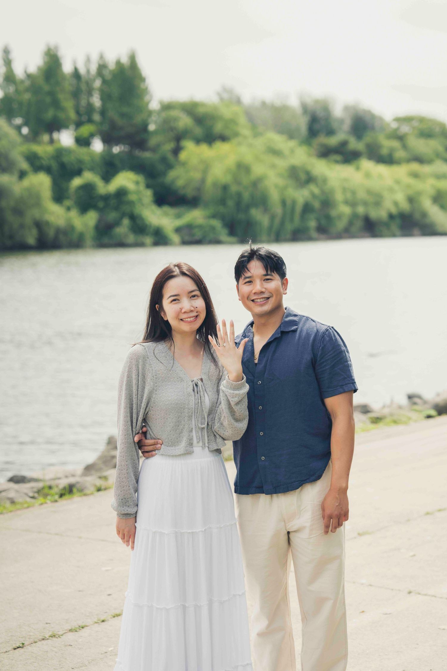 Happy couple posing by a lakeside, woman showing engagement ring, surrounded by lush green trees.