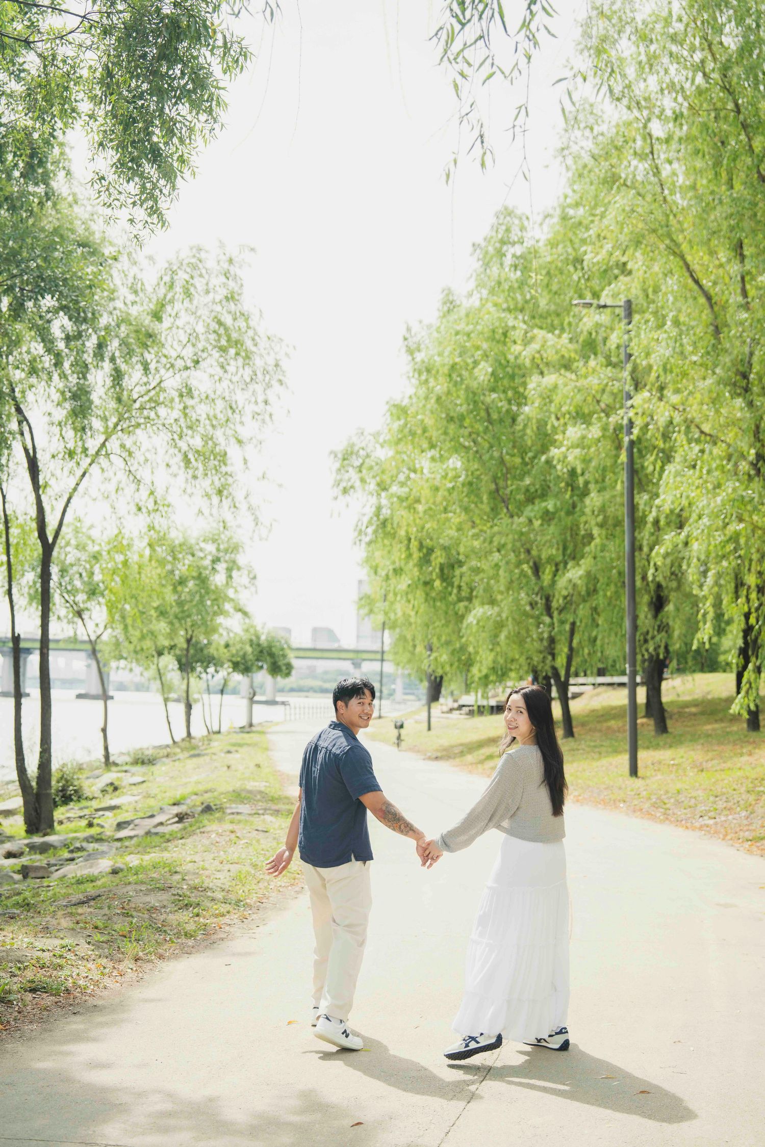 A couple holds hands on a scenic park path lined with willow trees near a river on a bright day.