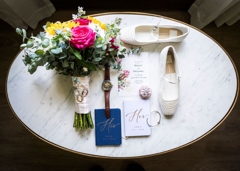 Wedding flat lay with colorful bridal bouquet, white shoes, rings, vow books, and invitation on marble table.