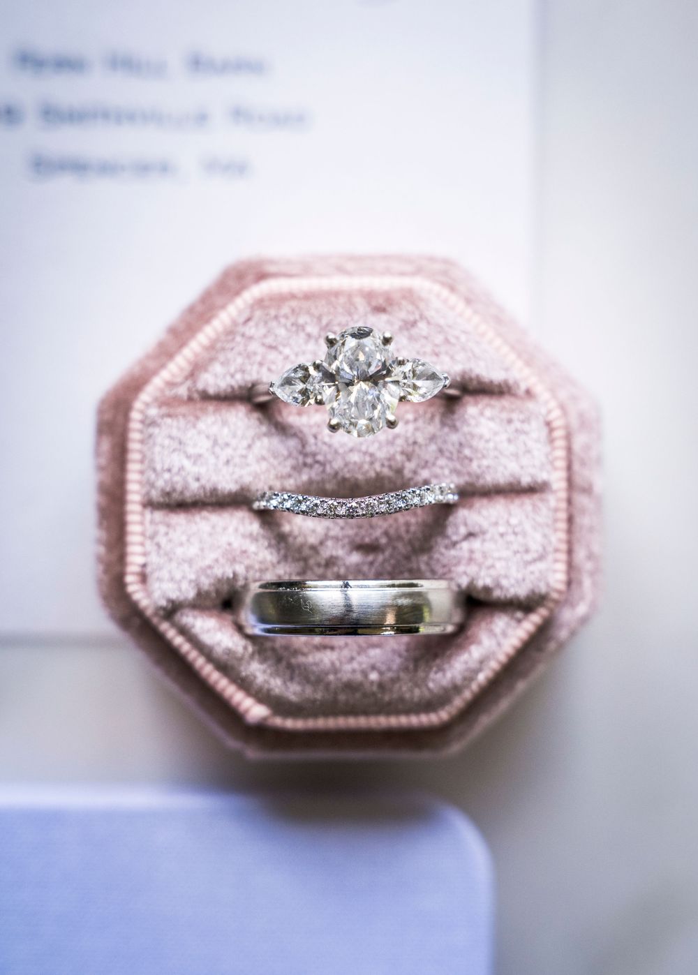 Three wedding rings displayed in a pink velvet octagonal ring box, featuring a diamond engagement ring, wedding band, and groom's ring.