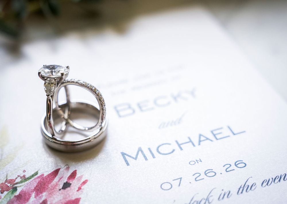 Diamond engagement ring and wedding band resting on Becky and Michael wedding invitation dated 07.26.26.