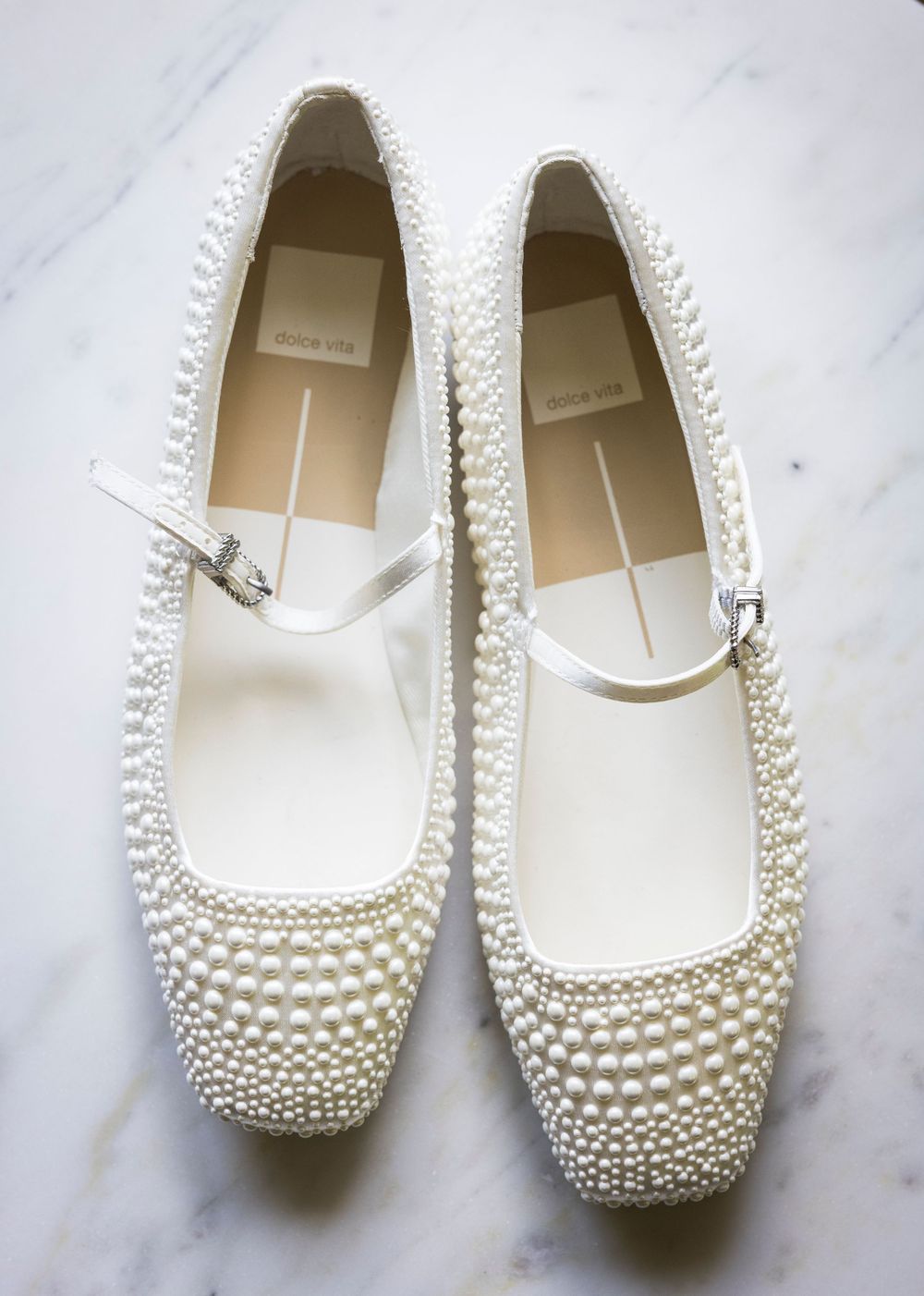White pearl-embellished Mary Jane ballet flats with ankle strap, displayed on marble surface.