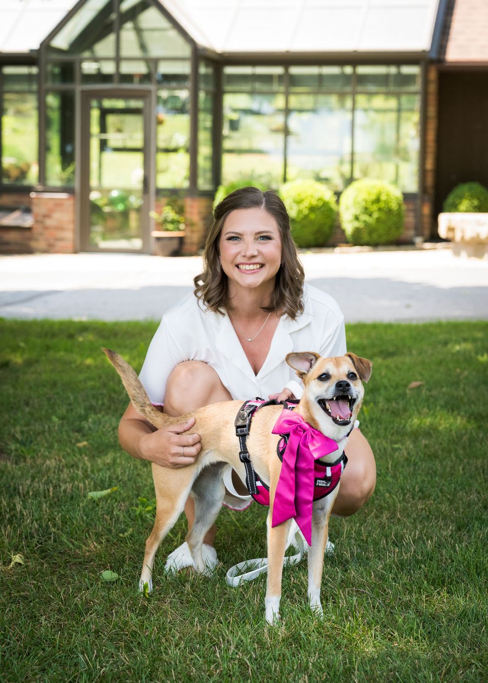 Smiling bride posing with her small dog wearing a pink bow on a green lawn outside a brick building.